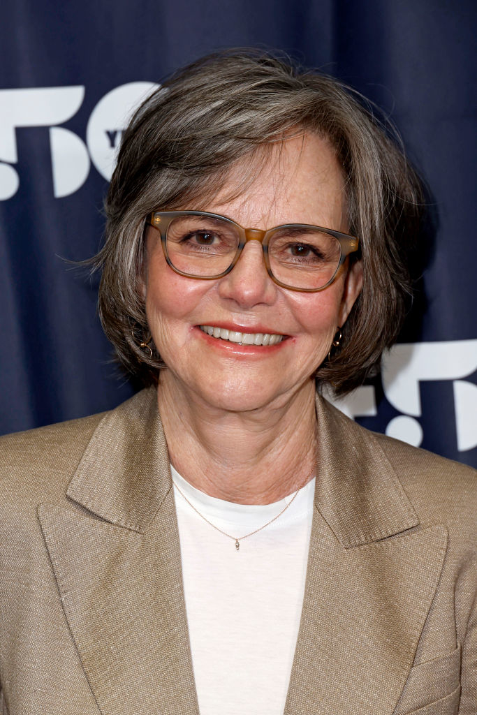 Sally Field smiles in a close-up photo at a press event, wearing glasses and a beige blazer over a white top