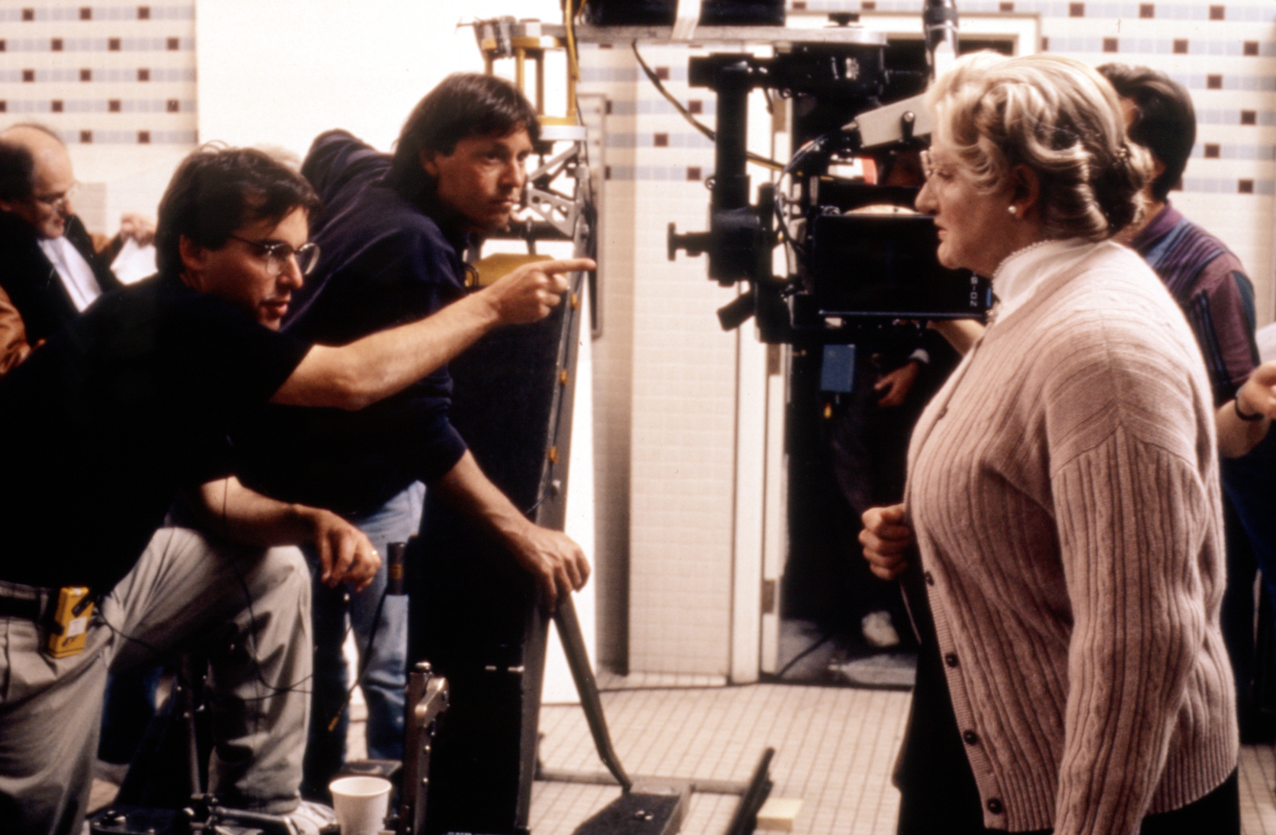 Robin Williams on set dressed as Mrs. Doubtfire, with film crew including director Chris Columbus in discussion, surrounded by movie filming equipment