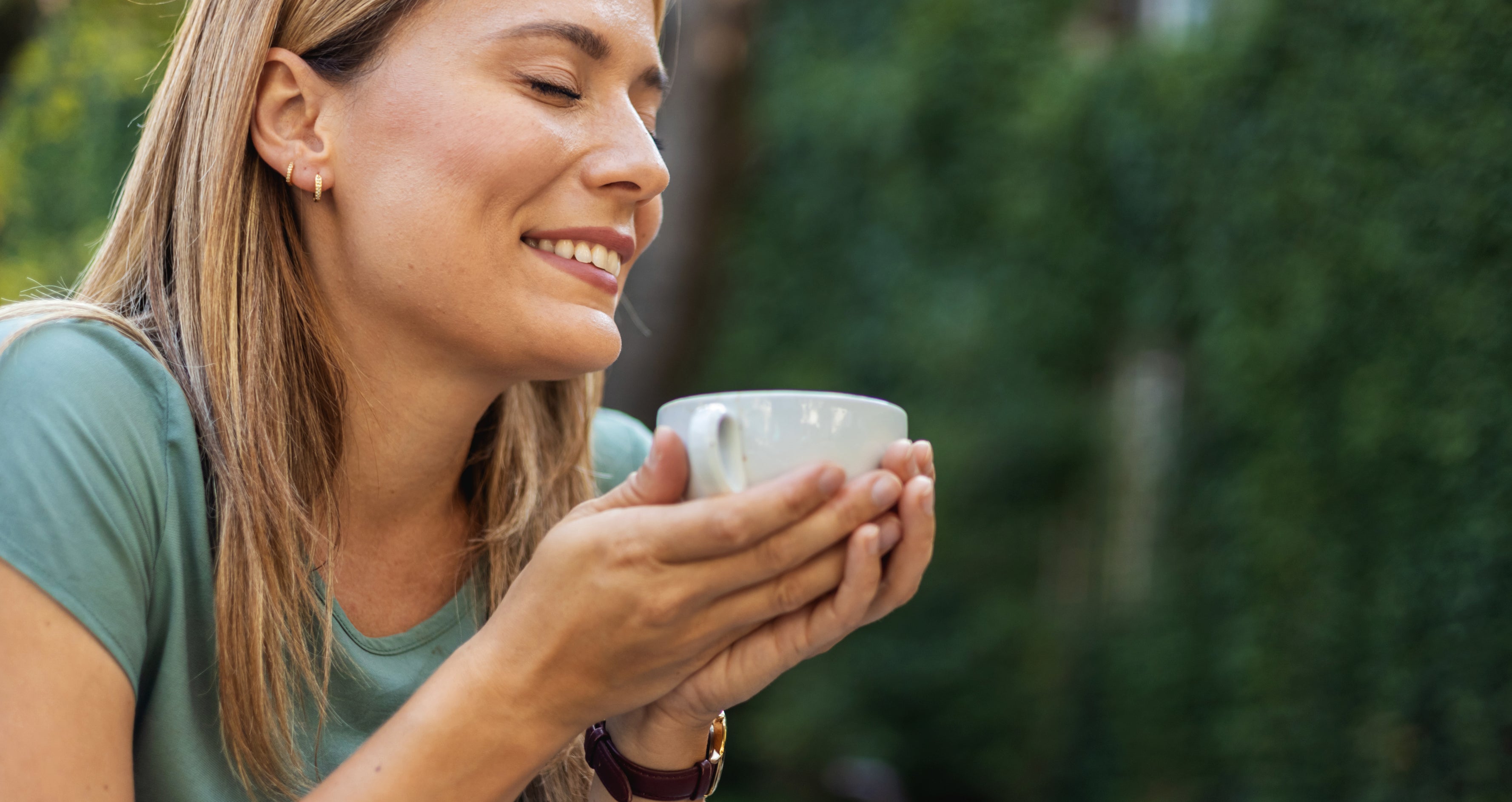 A woman with closed eyes holds a cup with both hands, appearing to enjoy the aroma. She is wearing a short-sleeved shirt, with trees and greenery in the background