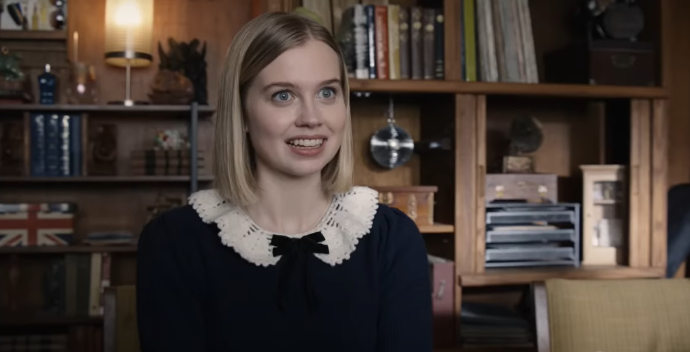Angourie Rice in a scene from Honor Society, smiles while sitting in a cozy, book-filled room. She wears a black dress with a white, ruffled collar