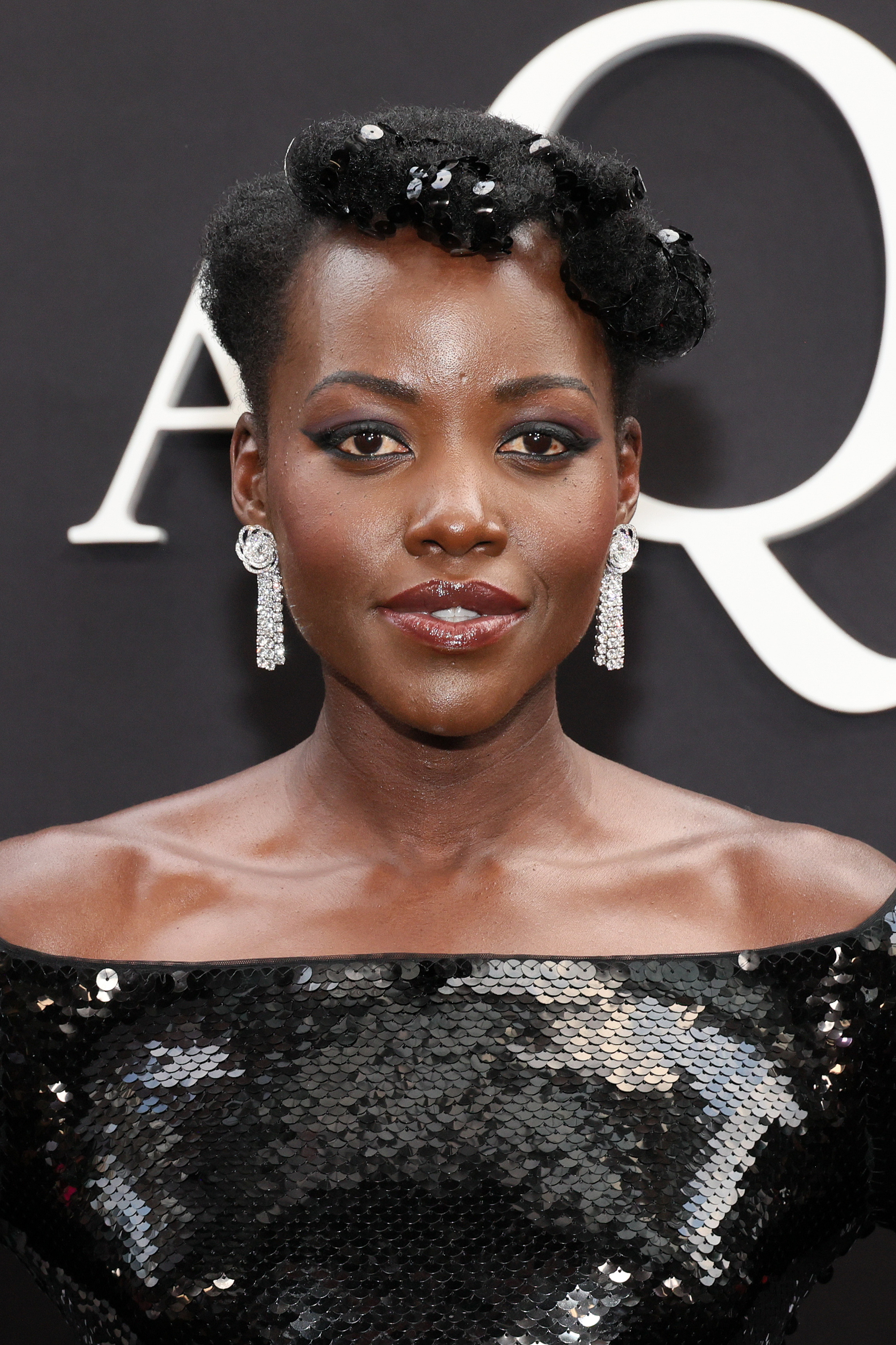 Lupita Nyong'o at a red carpet event, wearing an off-the-shoulder sequined black dress, with elegant earrings and styled hair