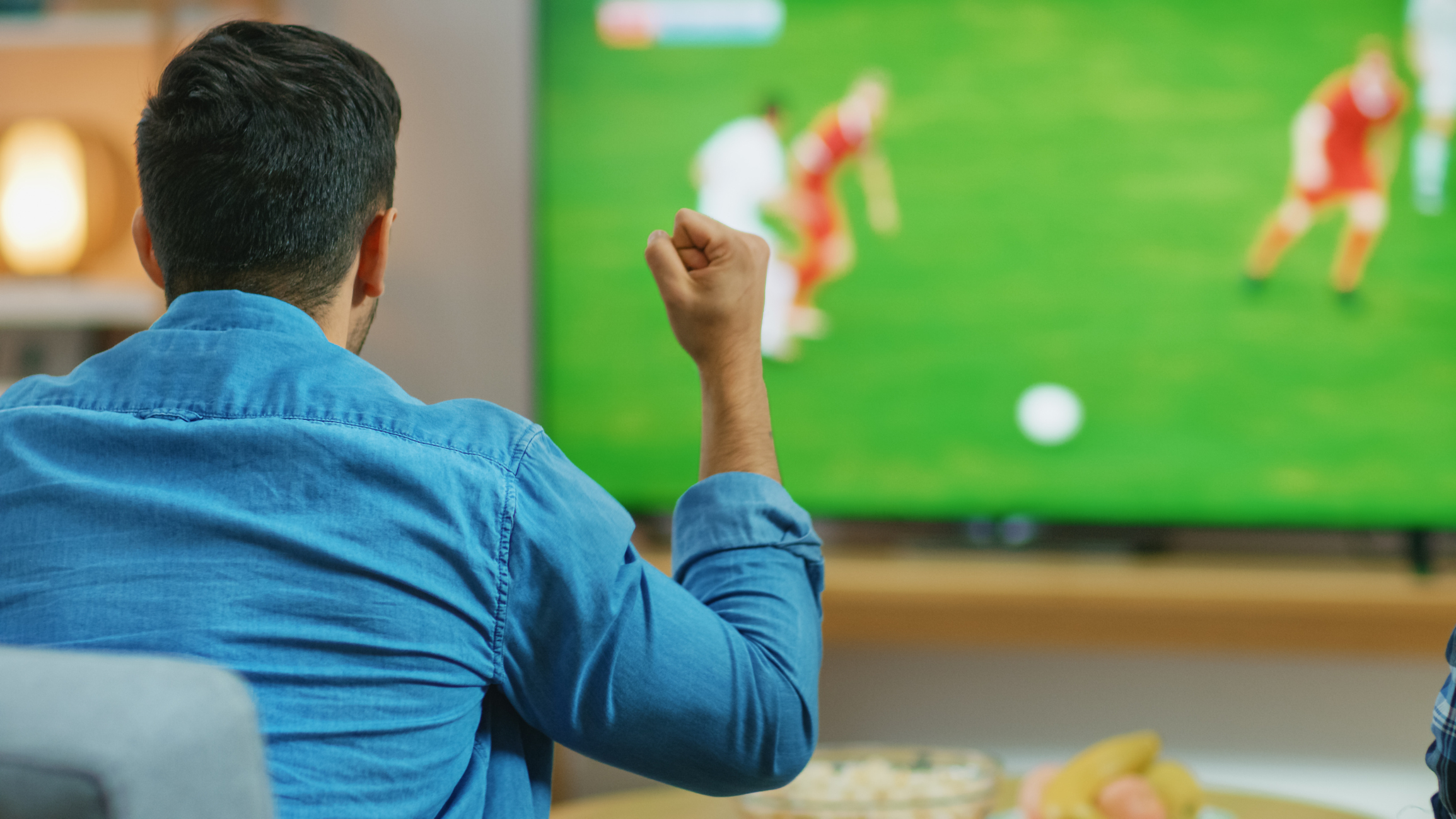 A person in a denim shirt watches a soccer match on TV with a raised fist, seemingly cheering while surrounded by snacks