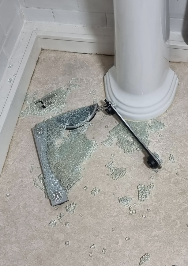 Broken glass fragments and a shattered mirror frame are scattered on the floor next to a white pedestal