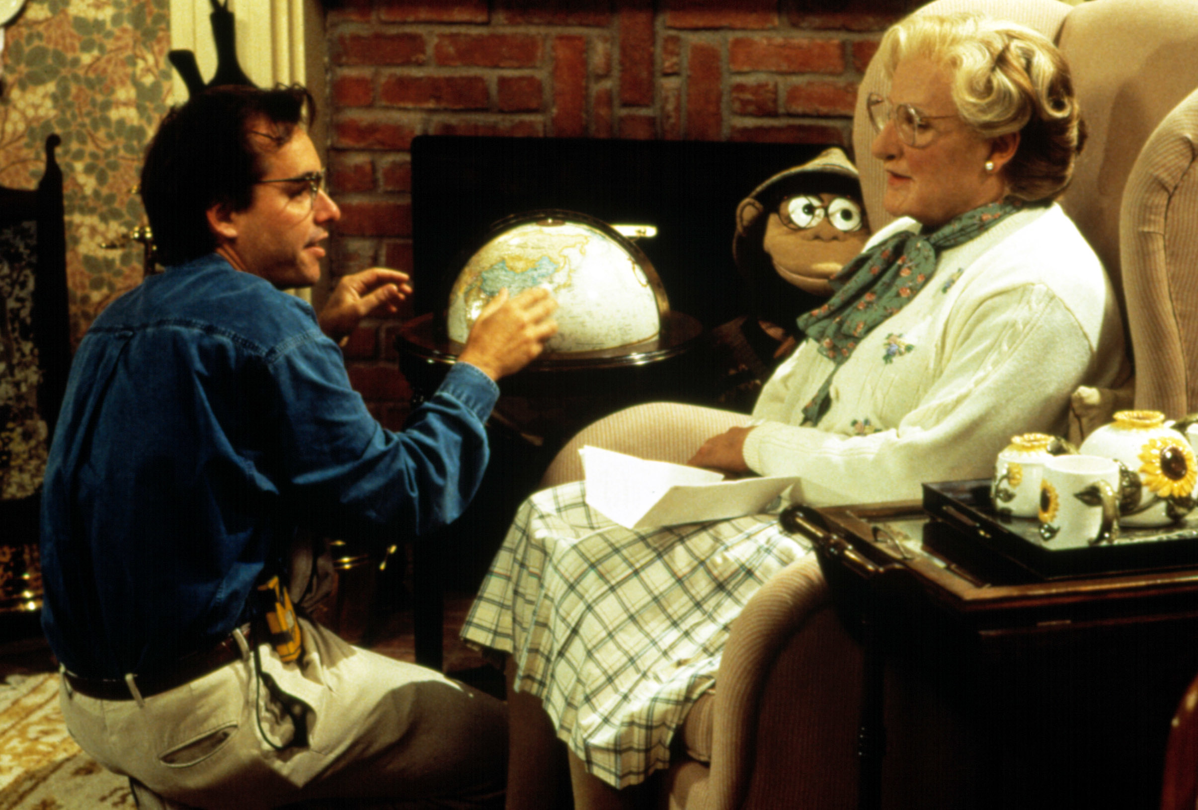 Robin Williams, as Mrs. Doubtfire, sits in a chair with a plaid skirt and white sweater, while Pierce Brosnan holds a globe, dressed in casual attire