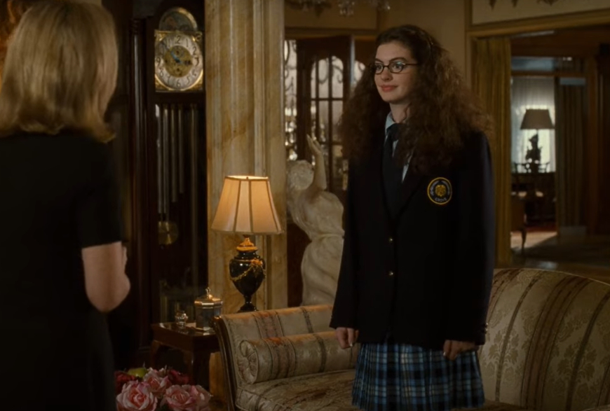 Anne Hathaway in crimped hair and wearing a school uniform with a blazer, standing in an elegantly furnished room, speaking with another person
