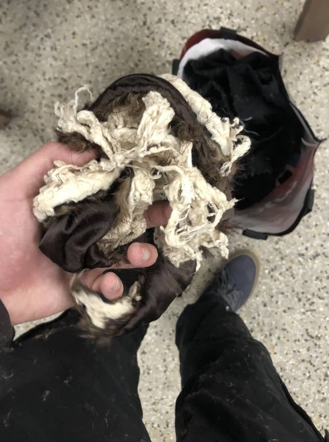 A person holds a torn, knotted piece of fabric above a black bag. The floor is speckled and unidentifiable