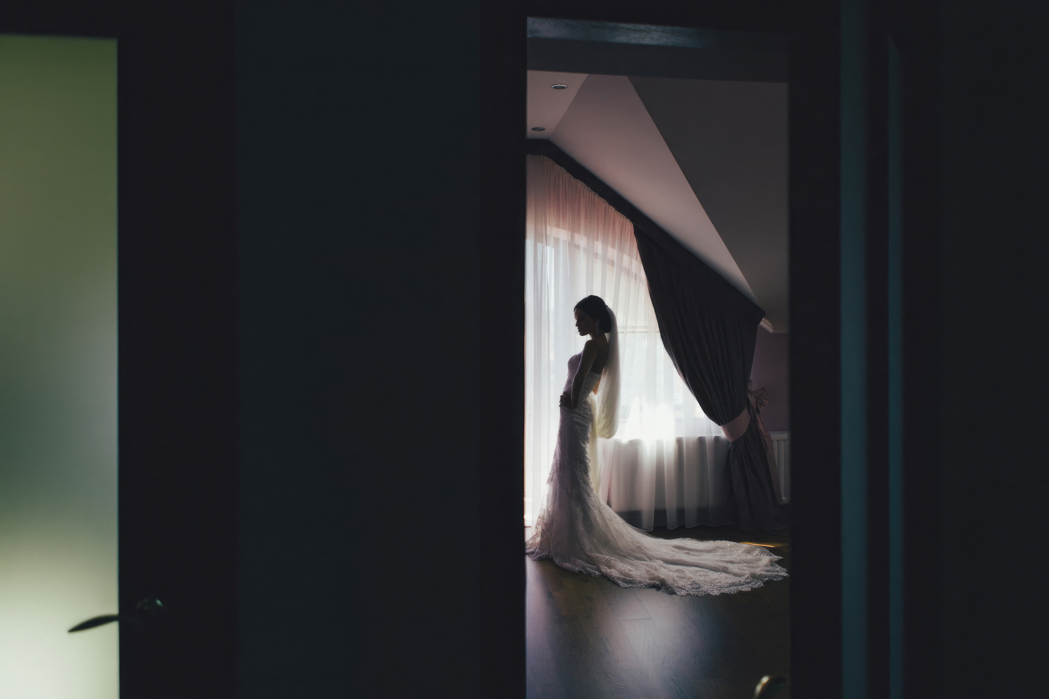 A bride stands alone  in front of a window wearing a long, elegant wedding dress with a train, partially hidden by shadows
