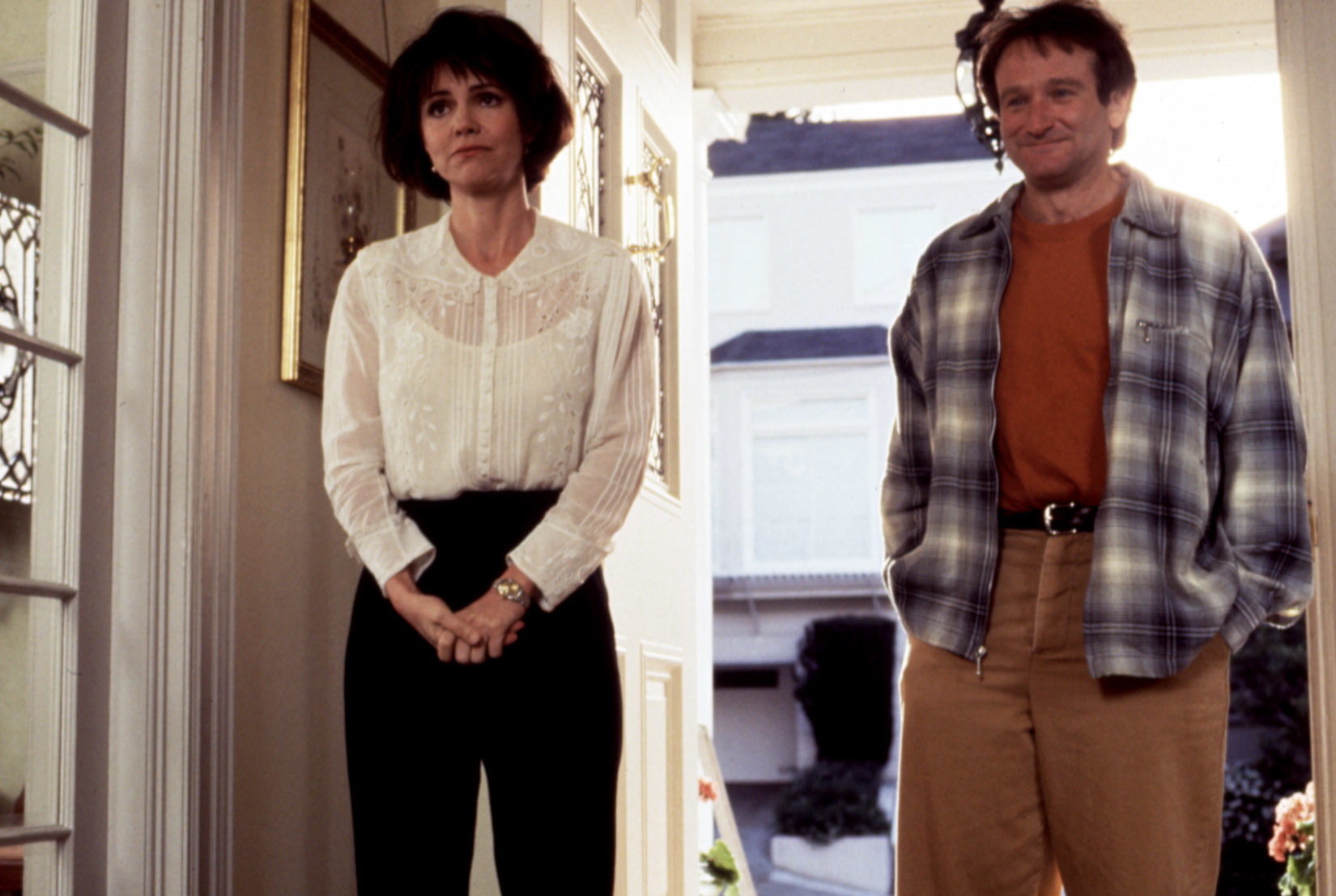 Sally Field and Robin Williams stand in a doorway, Sally in a white blouse and black pants, Robin in a plaid shirt over an orange top and beige pants