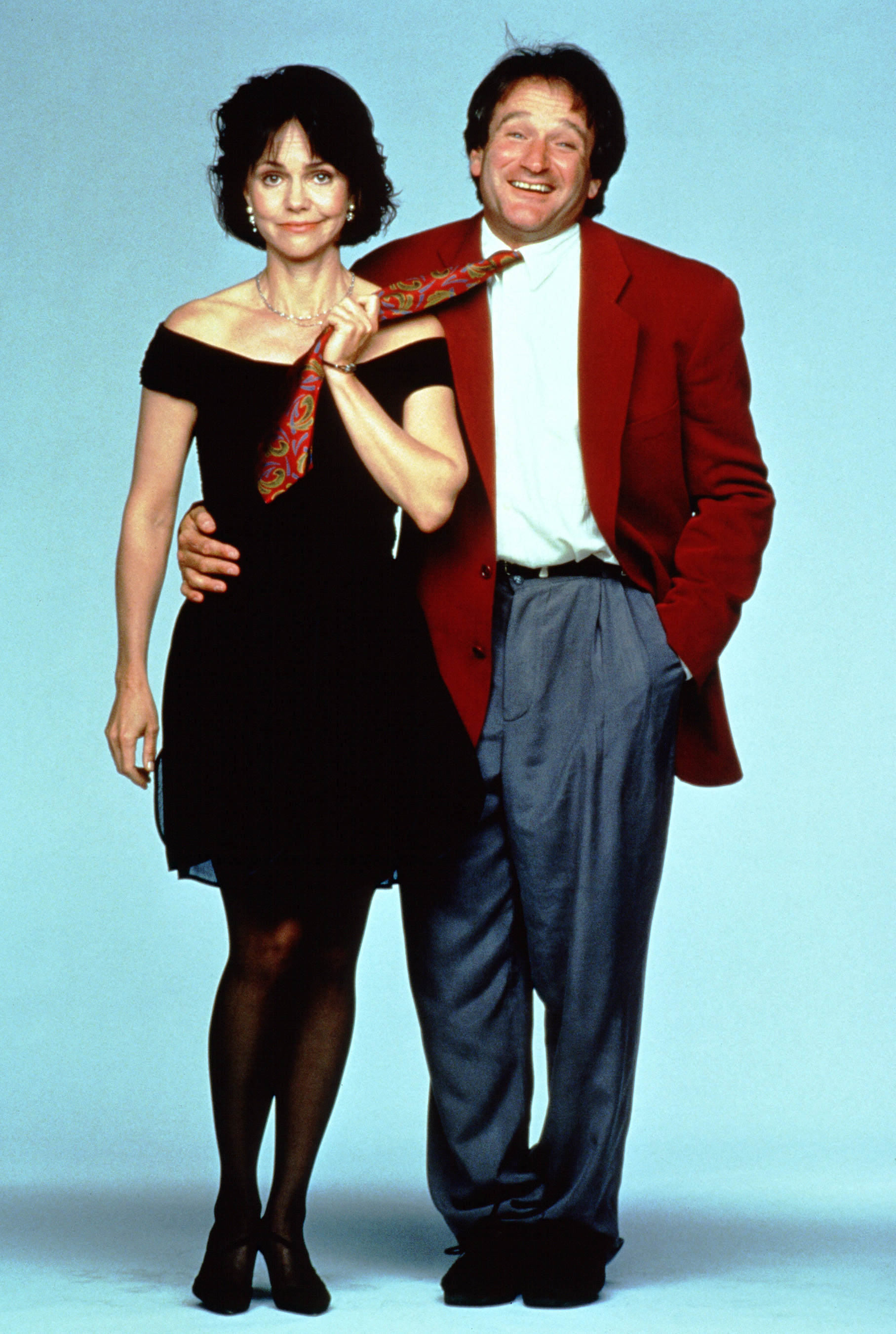 Sally Field and Robin Williams pose together; Williams is playfully holding a colorful tie around Field. She wears a black dress, and he wears a red blazer with gray pants