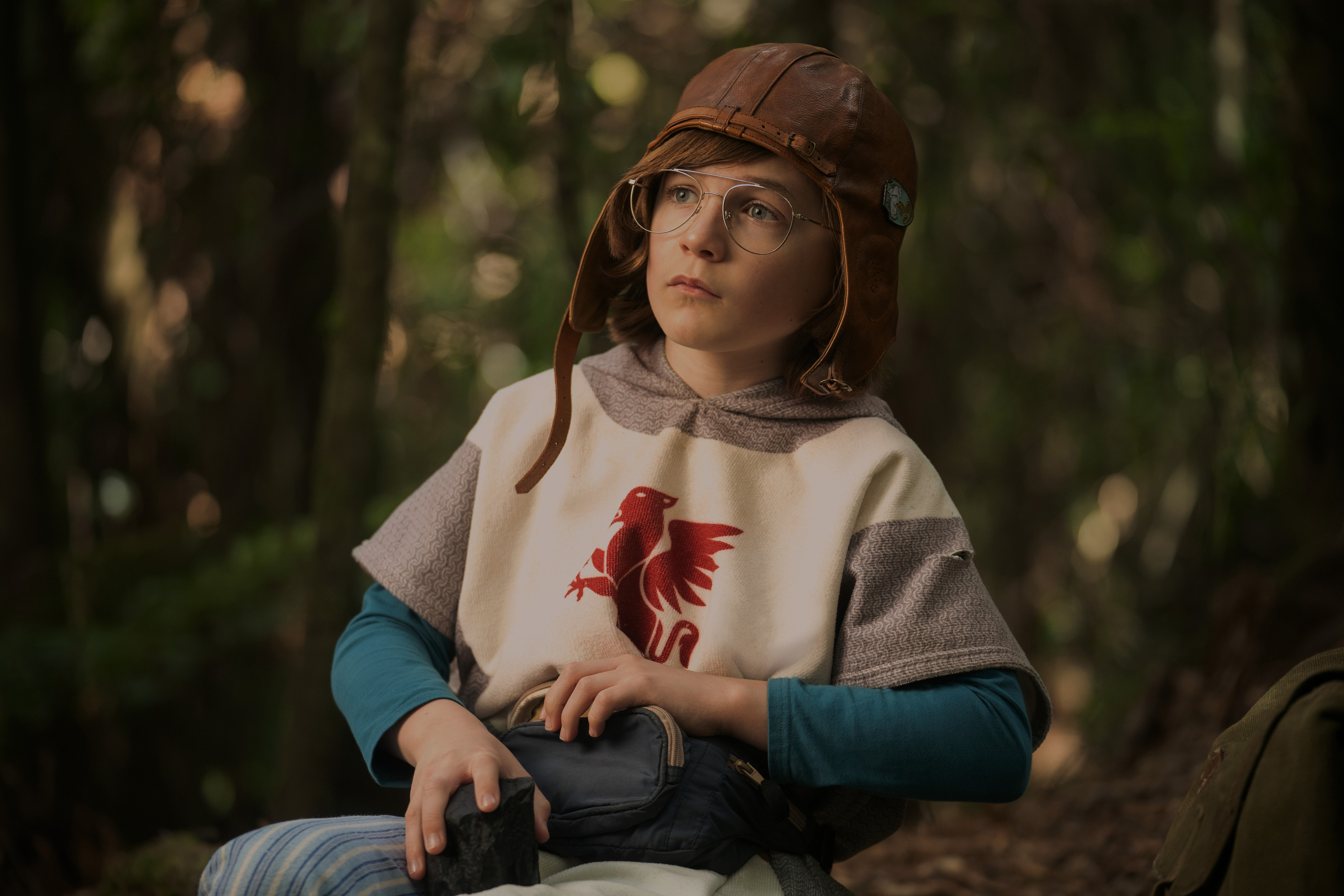 Young boy in a medieval-style tunic with a red dragon symbol, wearing a leather aviator cap and glasses, holding a small bag, in a forest setting