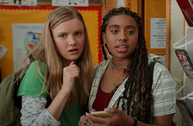 Two girls stand indoors, looking surprised. The girl on the left wears a green top with a blue long-sleeve shirt underneath, and the girl on the right wears a plaid shirt.