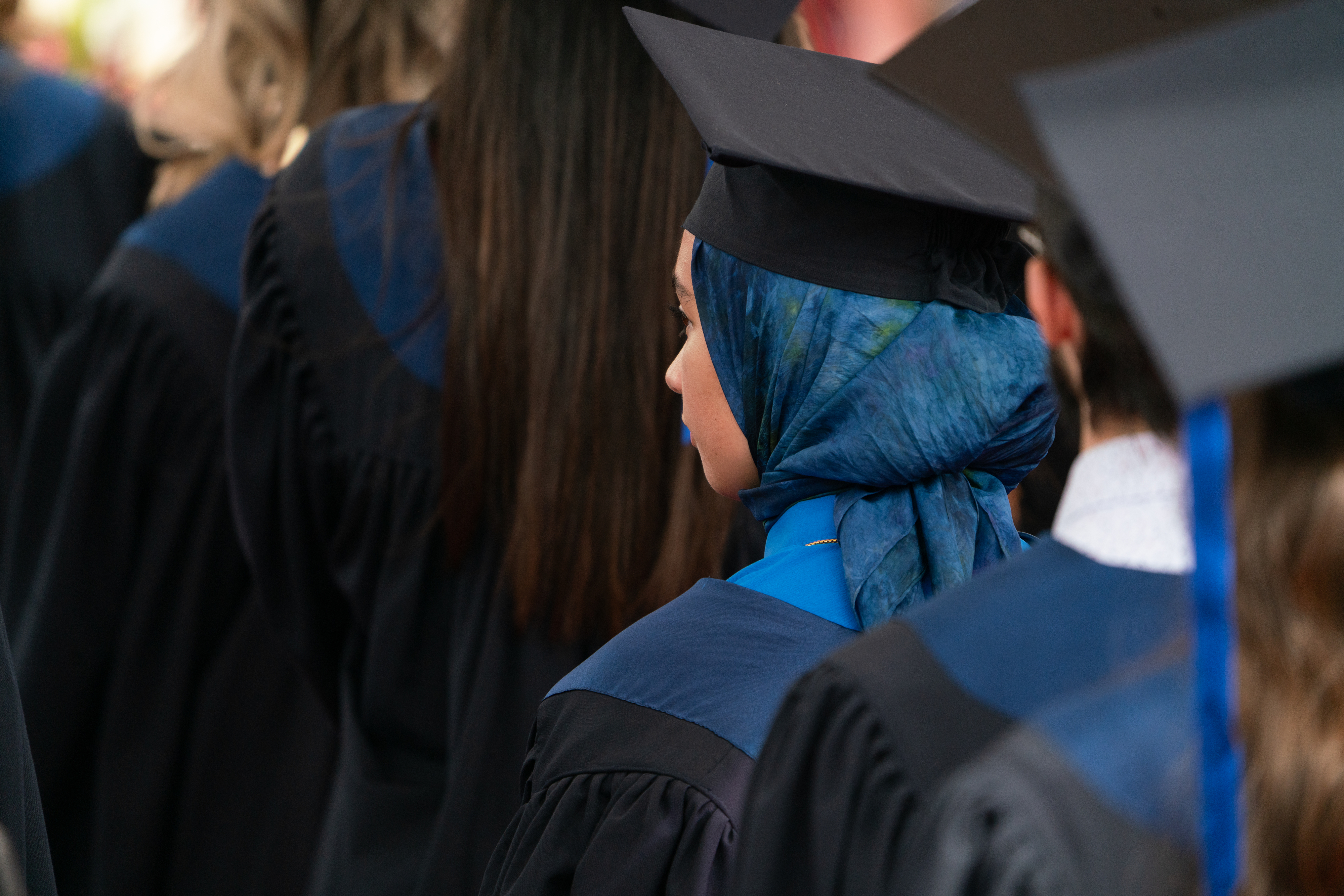 Graduates in caps and gowns stand in a line during a commencement ceremony. A person in a blue hijab is prominently shown among them
