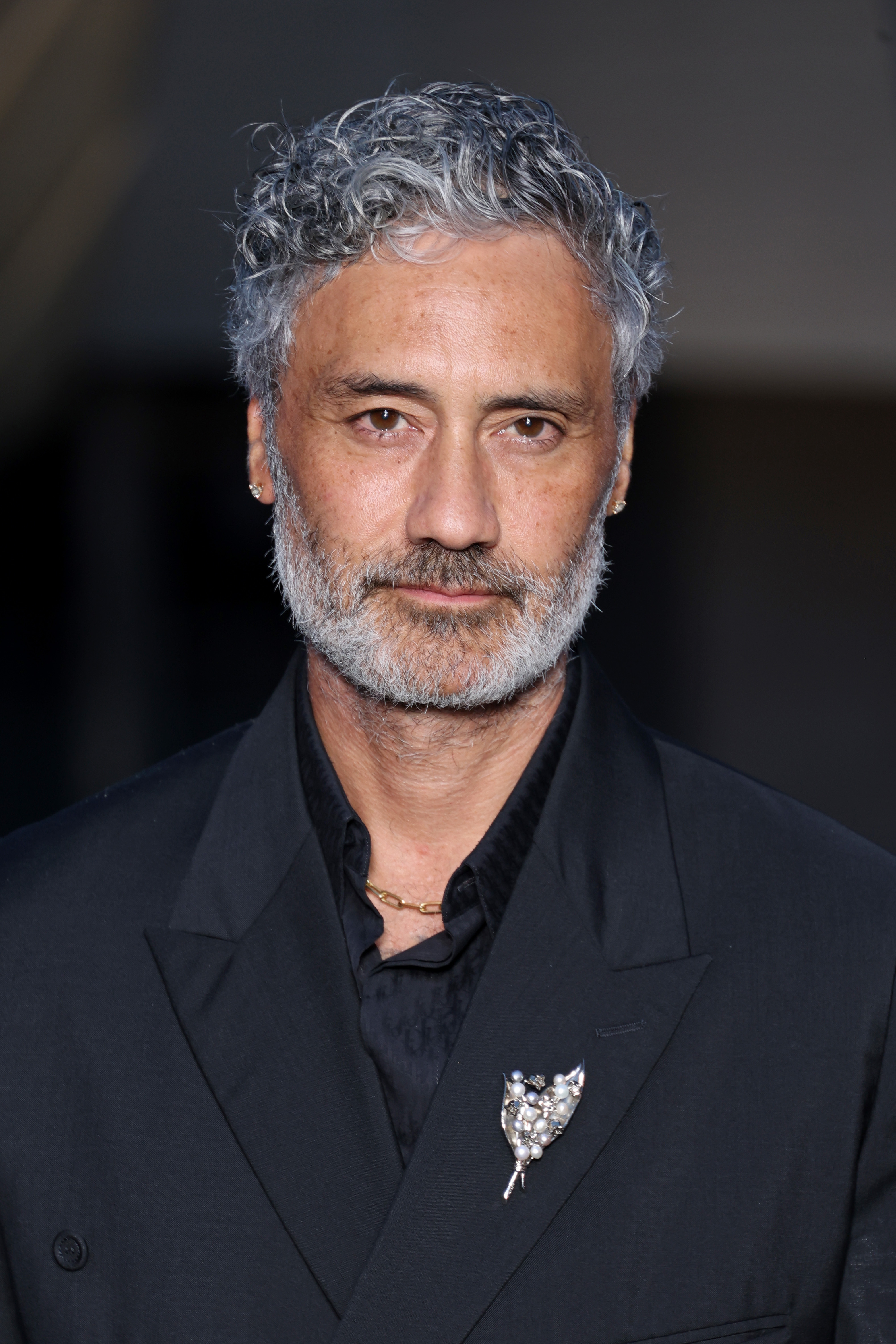 Taika Waititi in a dark suit with a decorative brooch, attends an event