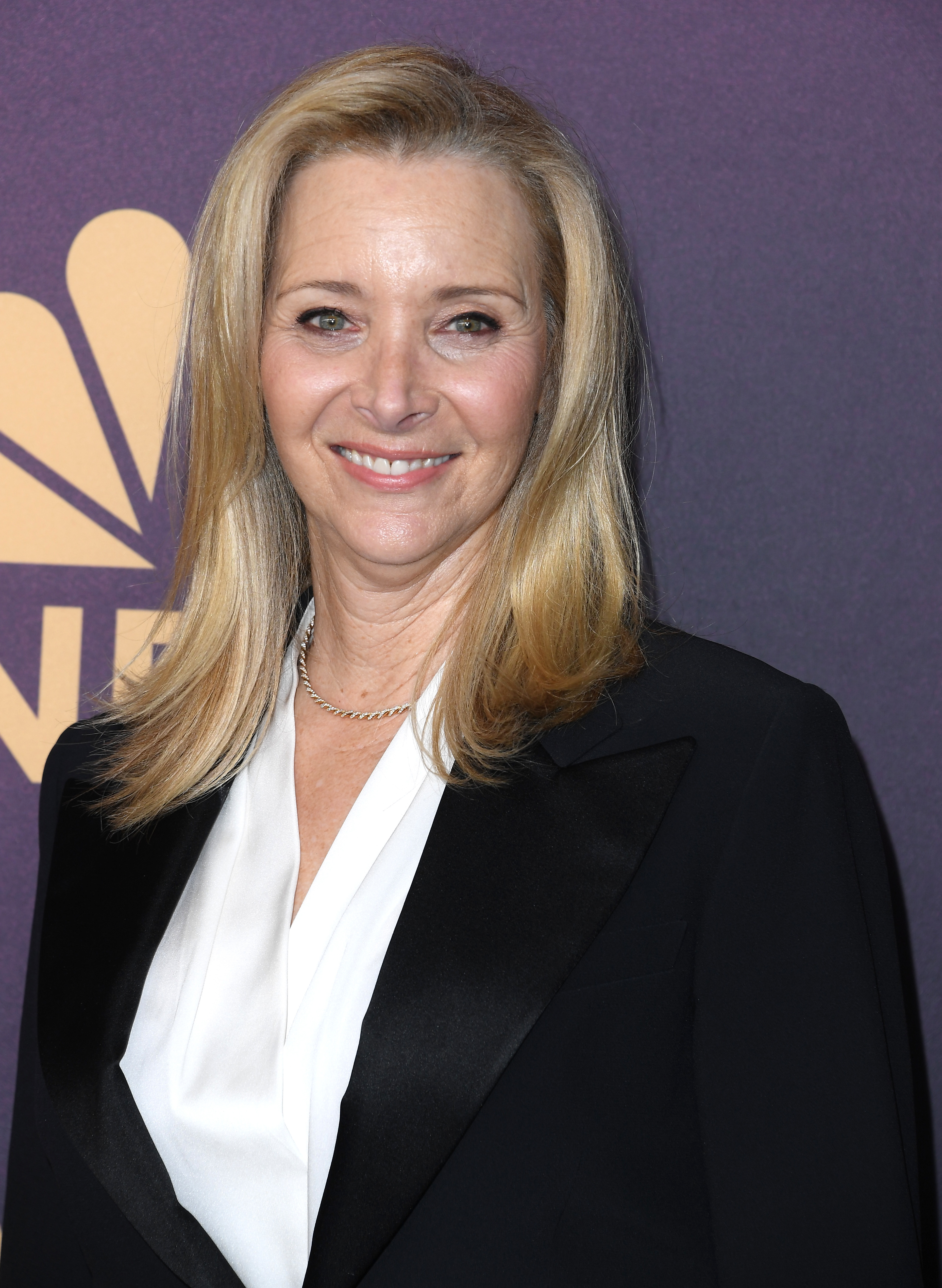 Lisa Kudrow smiles while wearing a formal black blazer over a white blouse at a media event