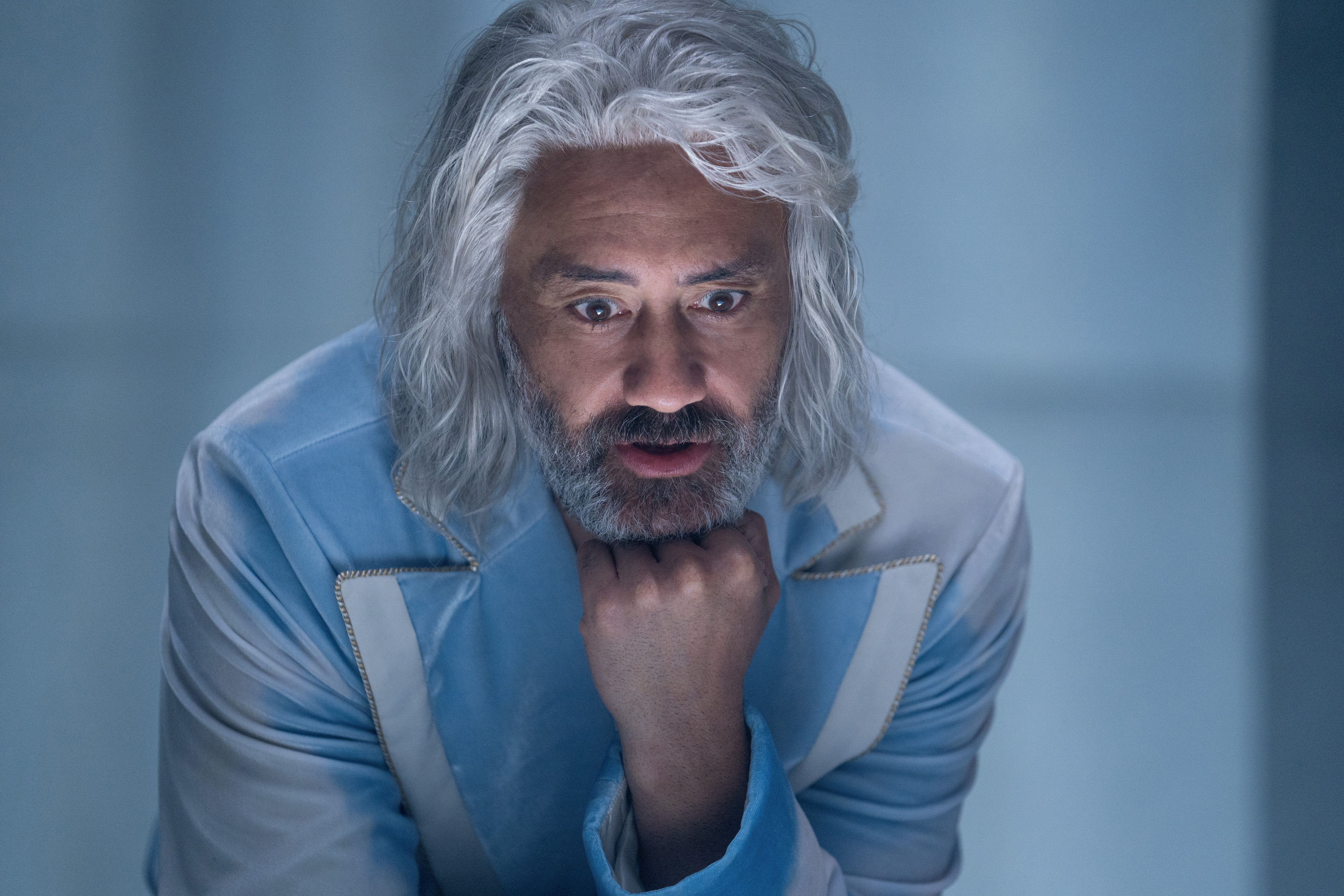 Taika Waititi, with long gray hair and a beard, wears a stylish light jacket and gazes downward with his chin resting on his fist