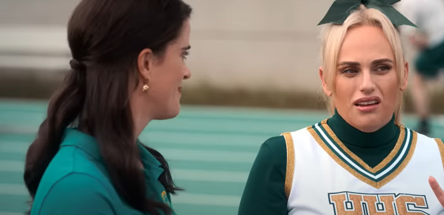Two women are talking; one wears a green polo shirt, while the other, Rebel Wilson, is dressed in a cheerleader uniform with "HHS" on it