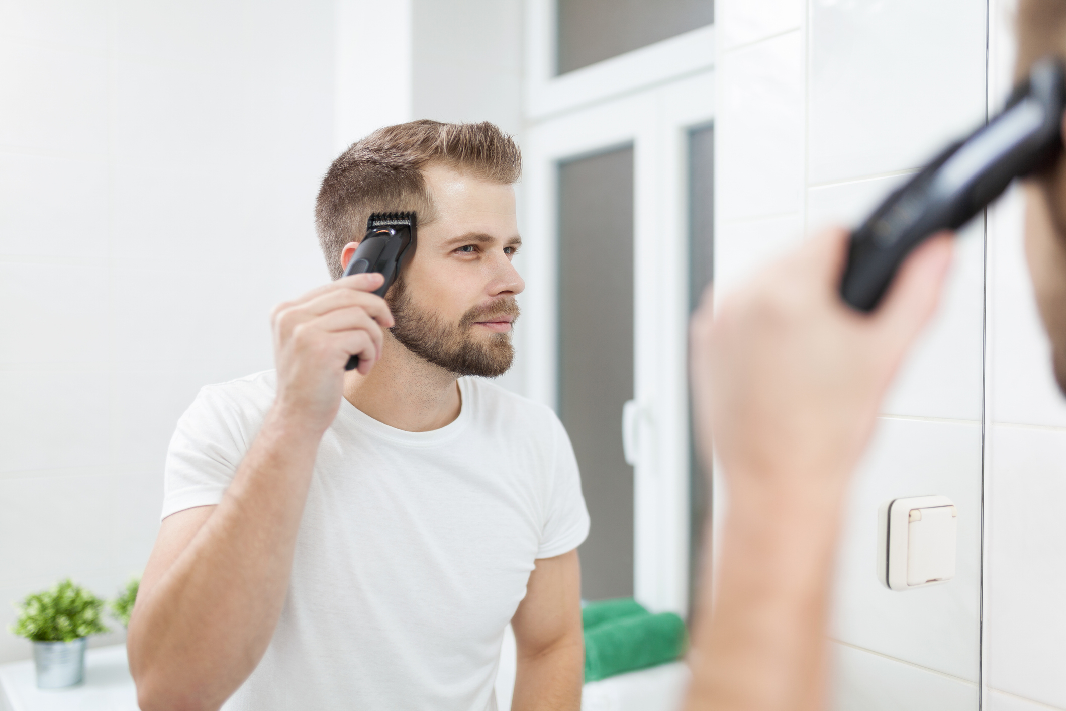 A man trims his hair using an electric clipper while looking in a bathroom mirror