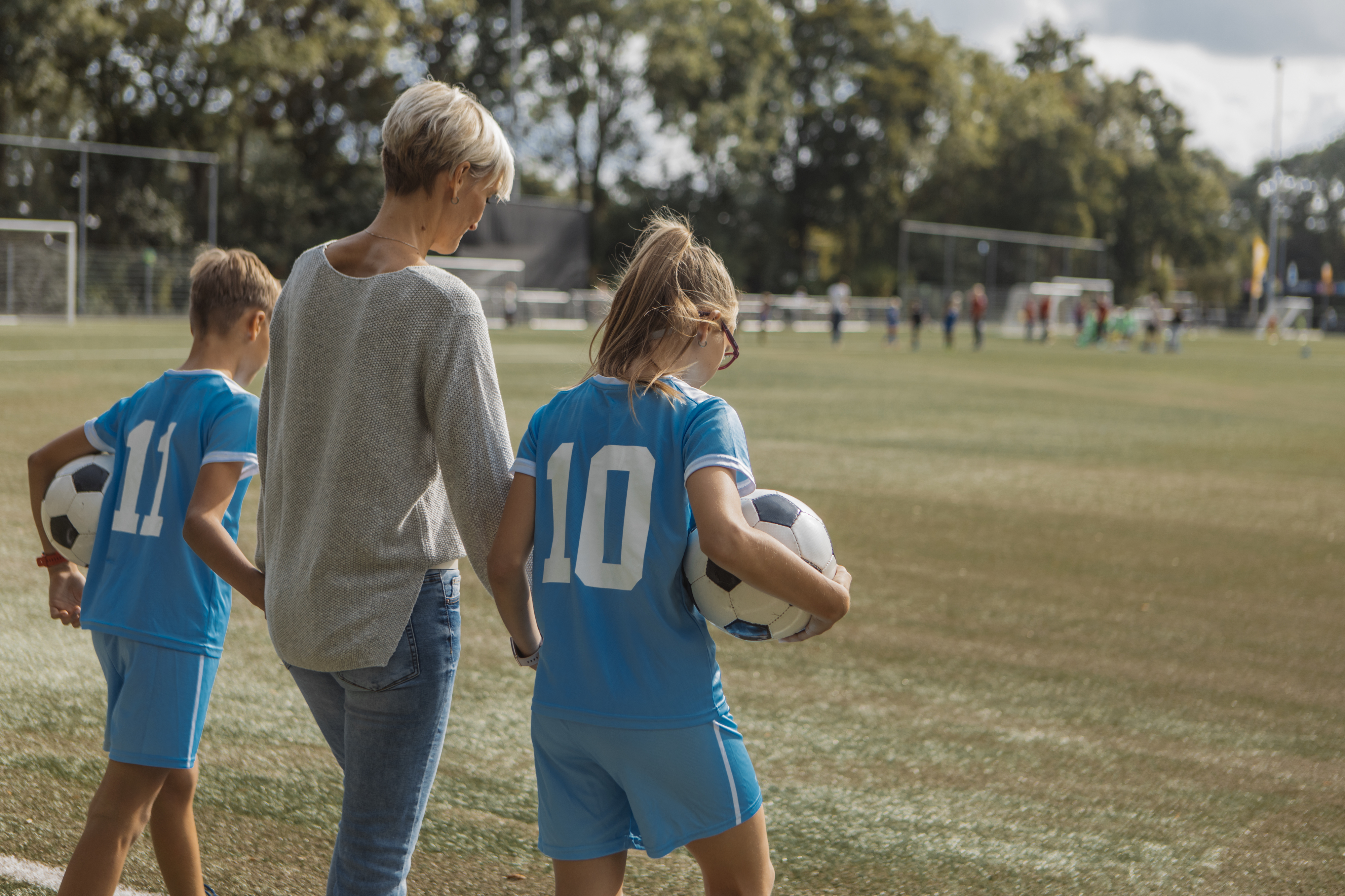 A woman walks with two young soccer players, wearing jerseys numbered 11 and 10, holding soccer balls on a soccer field