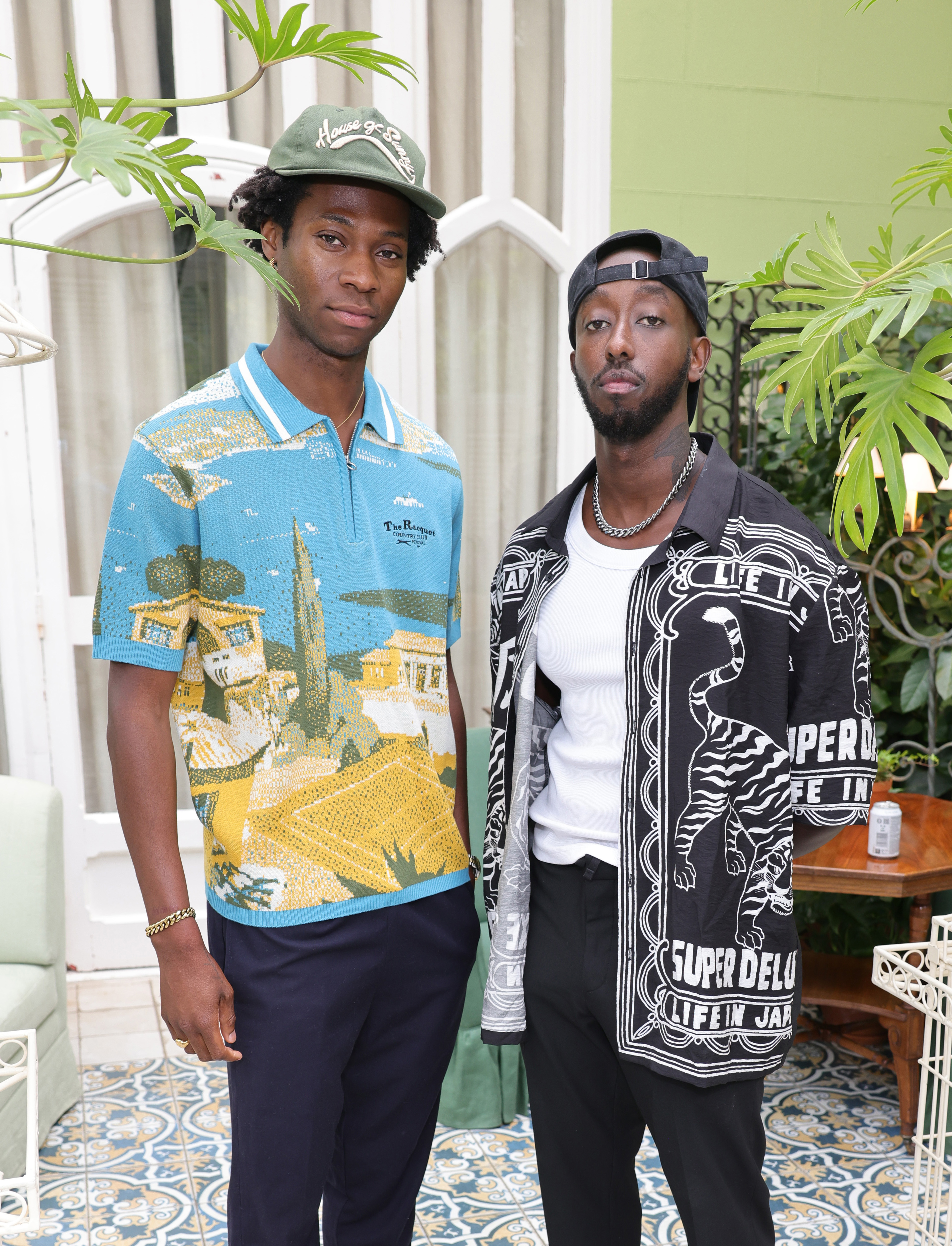 Two men pose together, one in a patterned polo shirt with an outdoor scene, and the other in a graphic button-up over a white tee, wearing a chain and cap