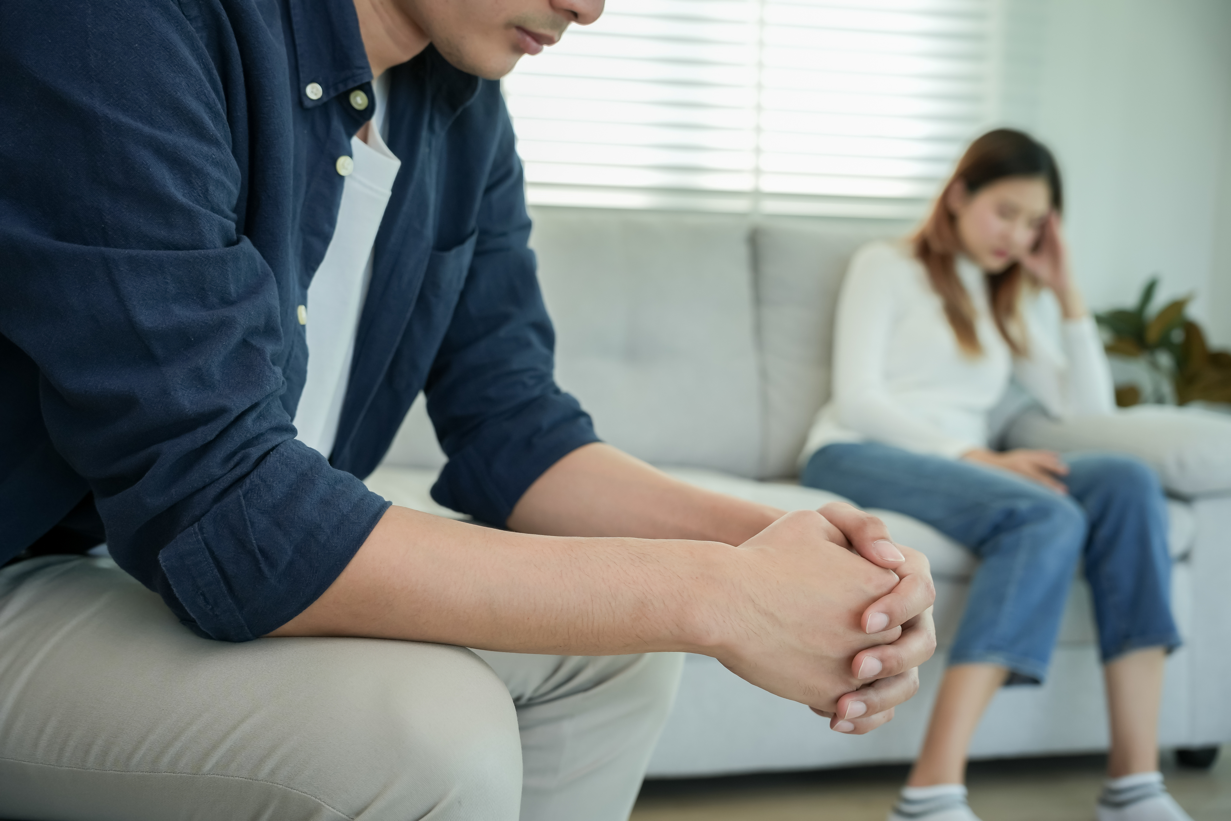 A man in a blue shirt and khaki pants sits on a couch with hands clasped, while a woman in a white sweater and jeans sits in the background, looking down