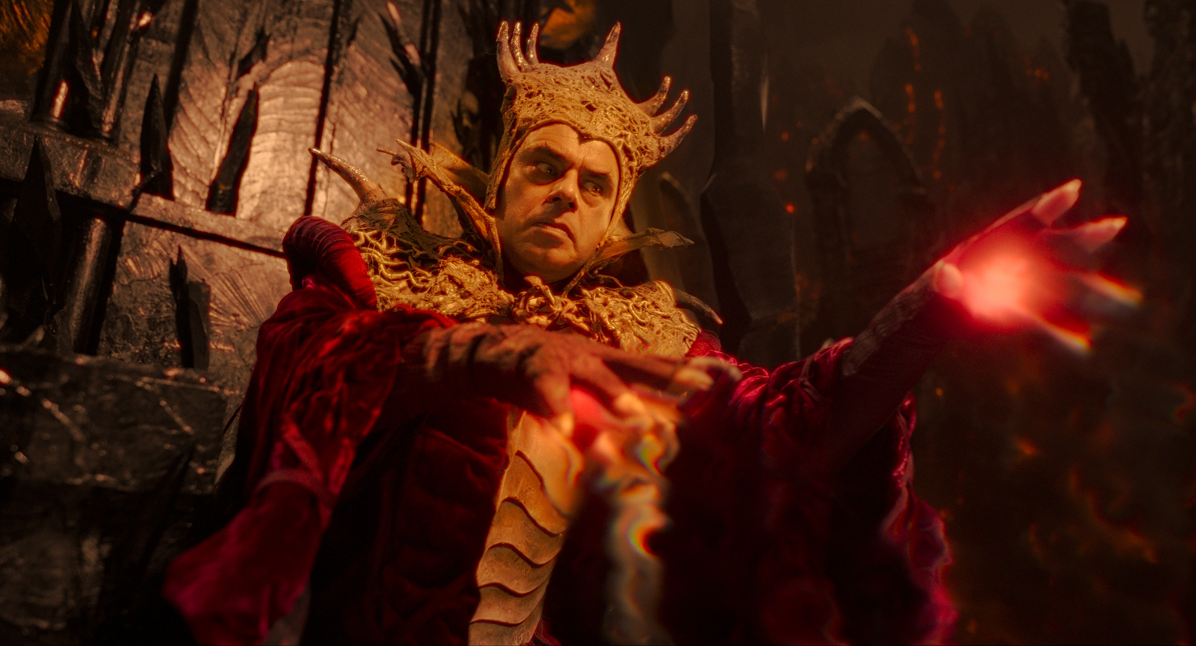 Jemaine Clement dressed extravagantly as a fantasy ruler with a golden crown and ornate armor, extends his glowing hand in a scene