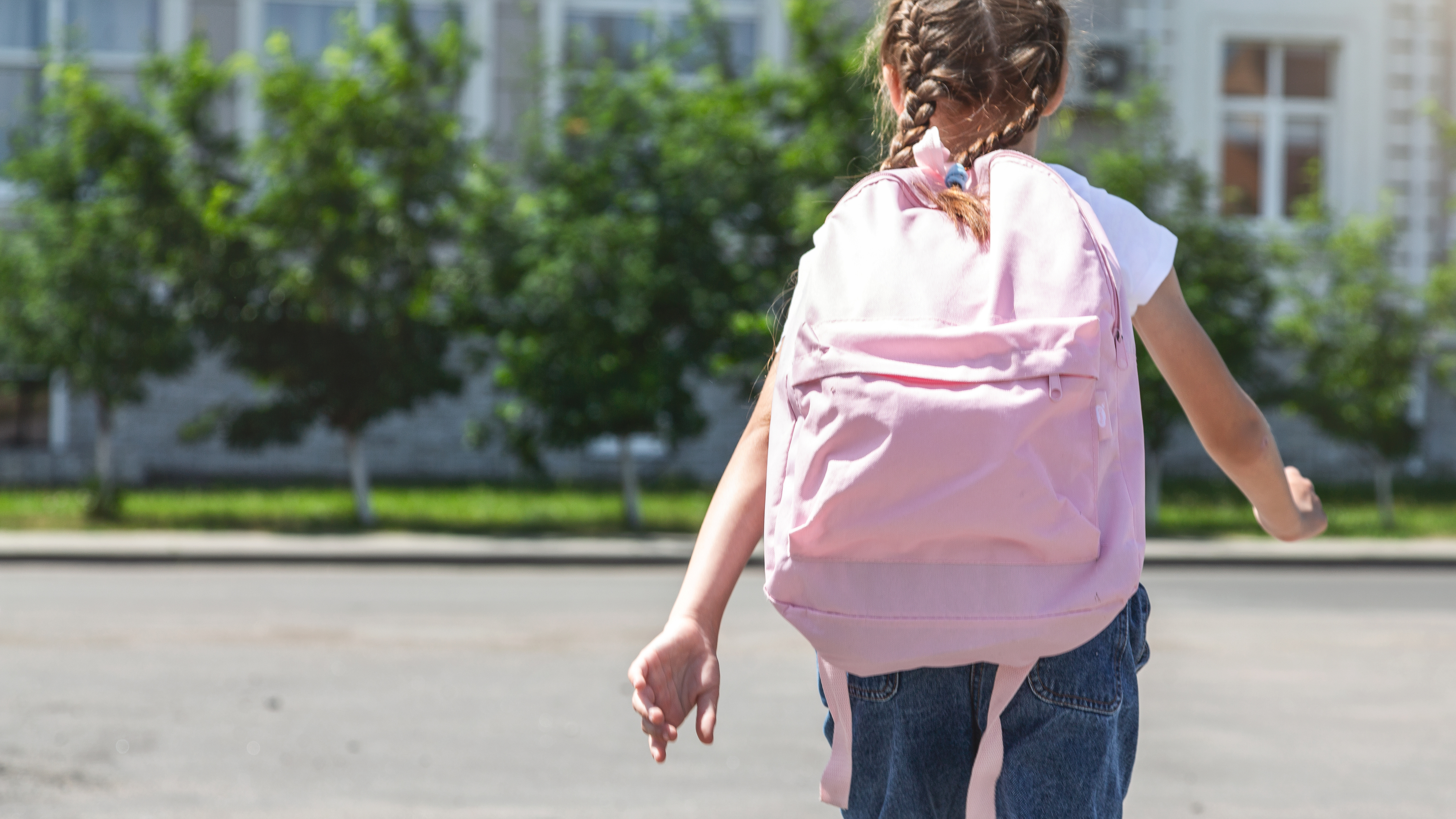 A young girl with braided hair is walking away with a large backpack on her shoulders, towards what appears to be a school building