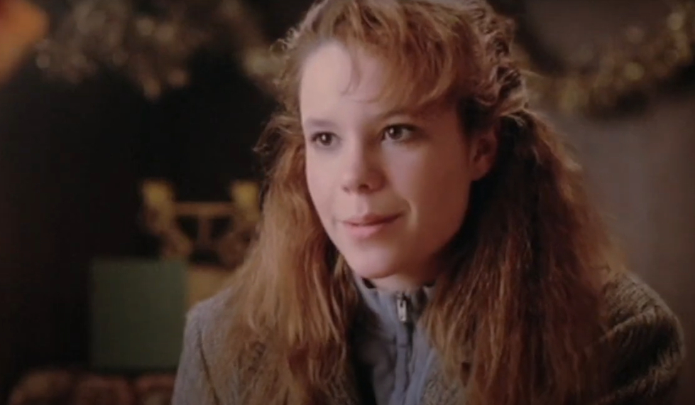Robyn Liverly with wavy hair smiles, wearing a zip-up jacket and a brown coat, in an indoor setting