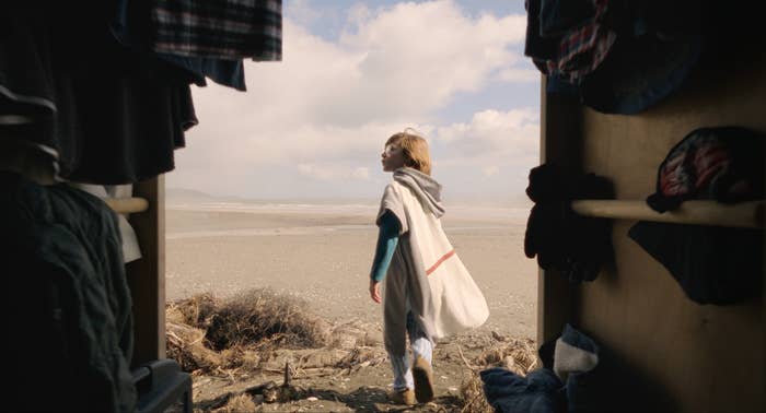 A child in a cape walks on a beach, viewed from inside a tent or similar shelter with hanging clothes. Sky and sea are in the background