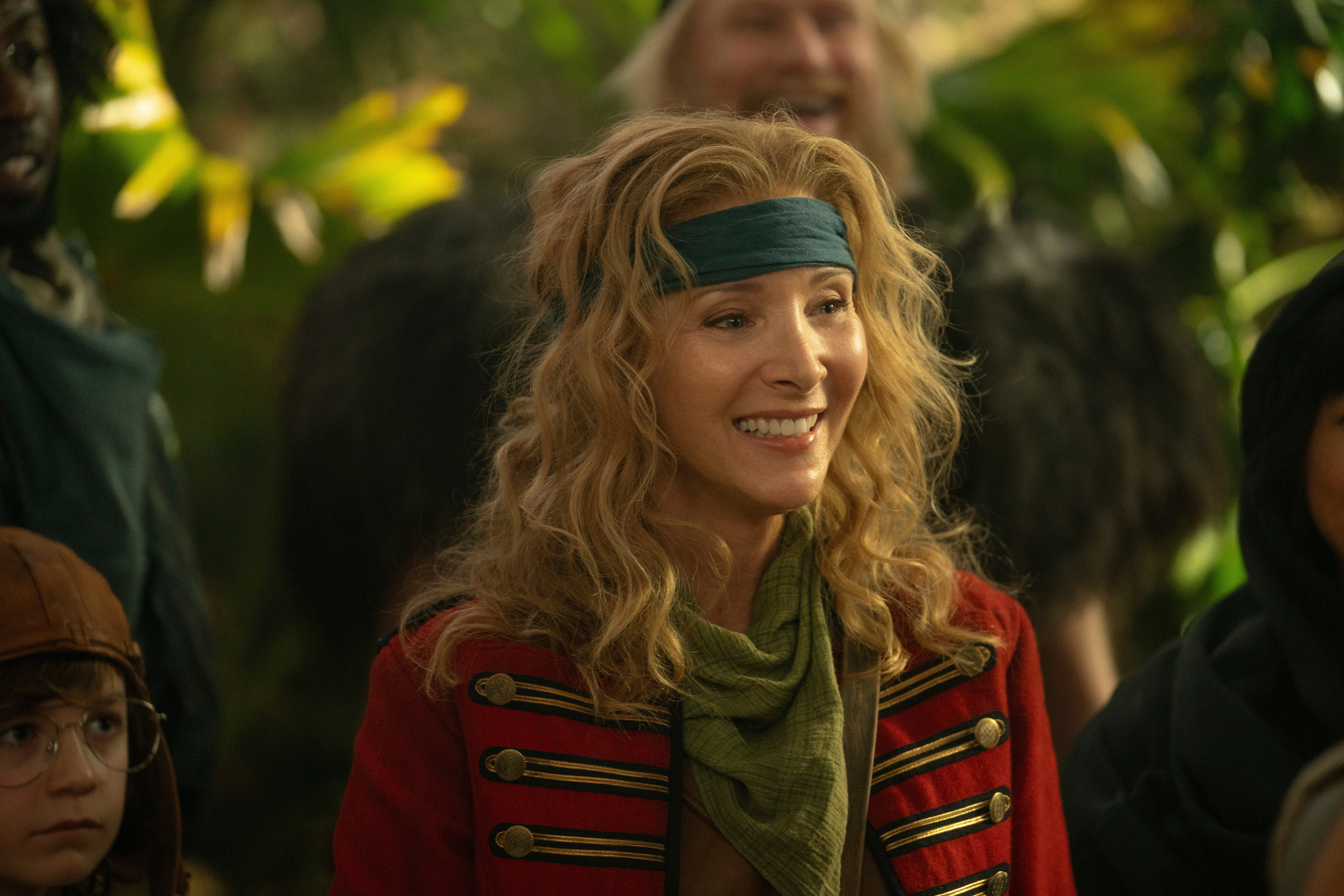 Lisa Kudrow in an adventurous scene wearing a red military-style jacket, green scarf, and headband, surrounded by other characters in rustic attire