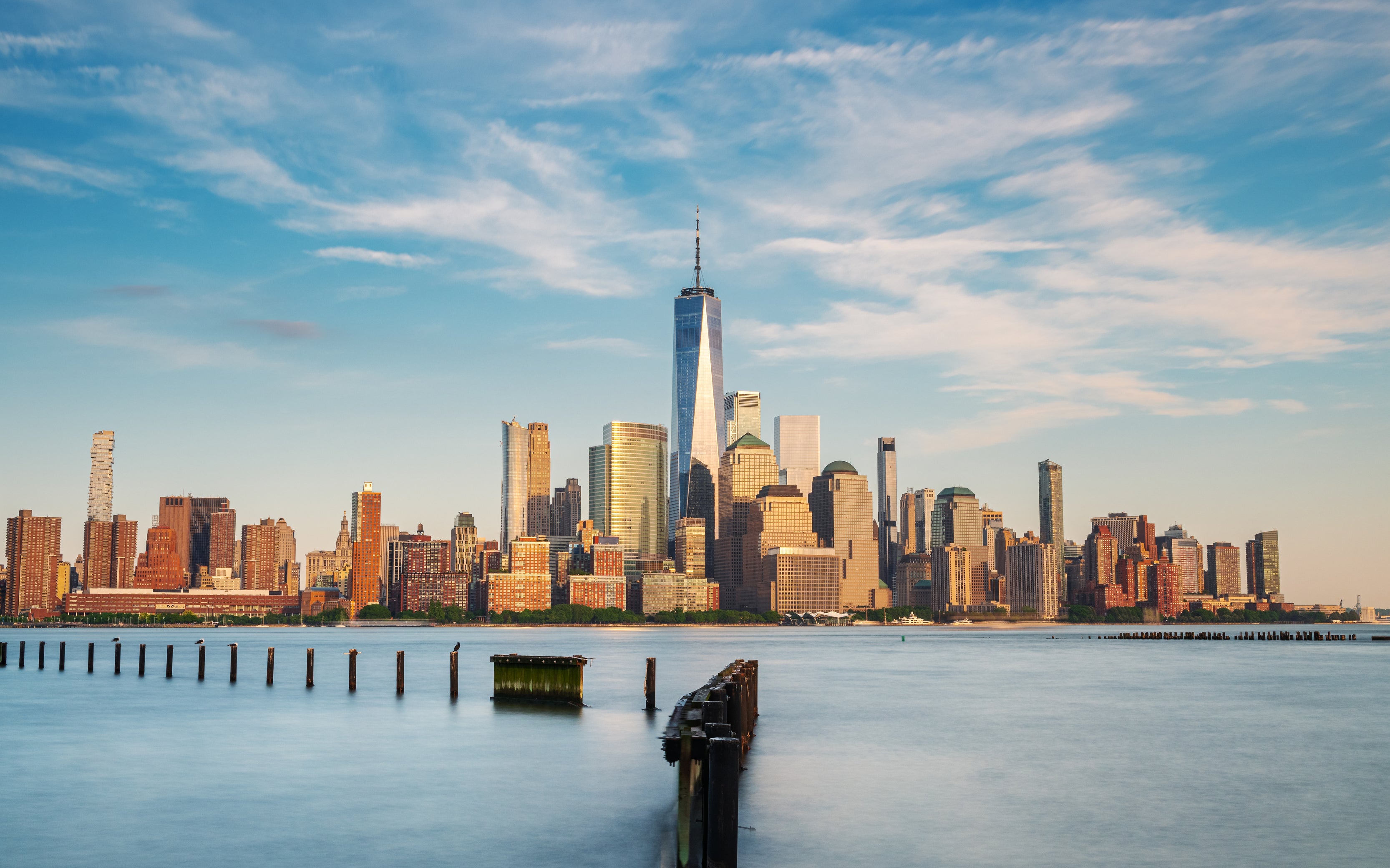 Skyline view of New York City with One World Trade Center prominently in the center, taken from the water with a sunny, blue sky backdrop