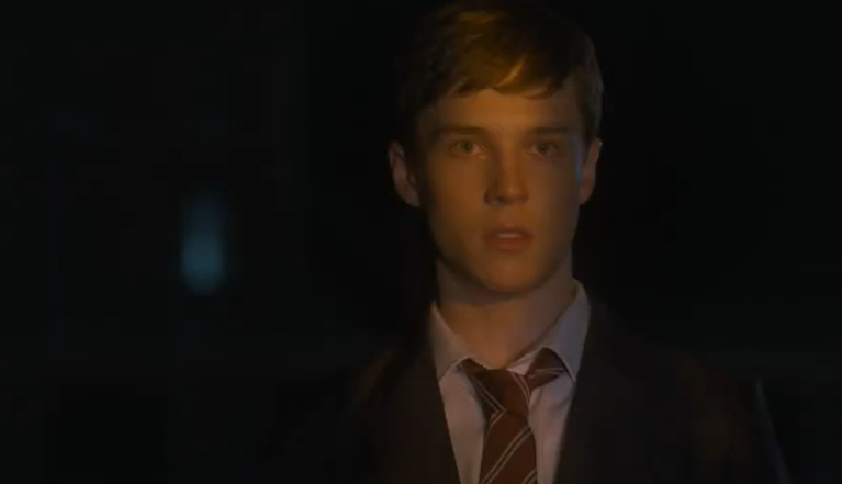 A young man with short brown hair, in a suit and tie, is shown in close-up, looking ahead with a serious expression.