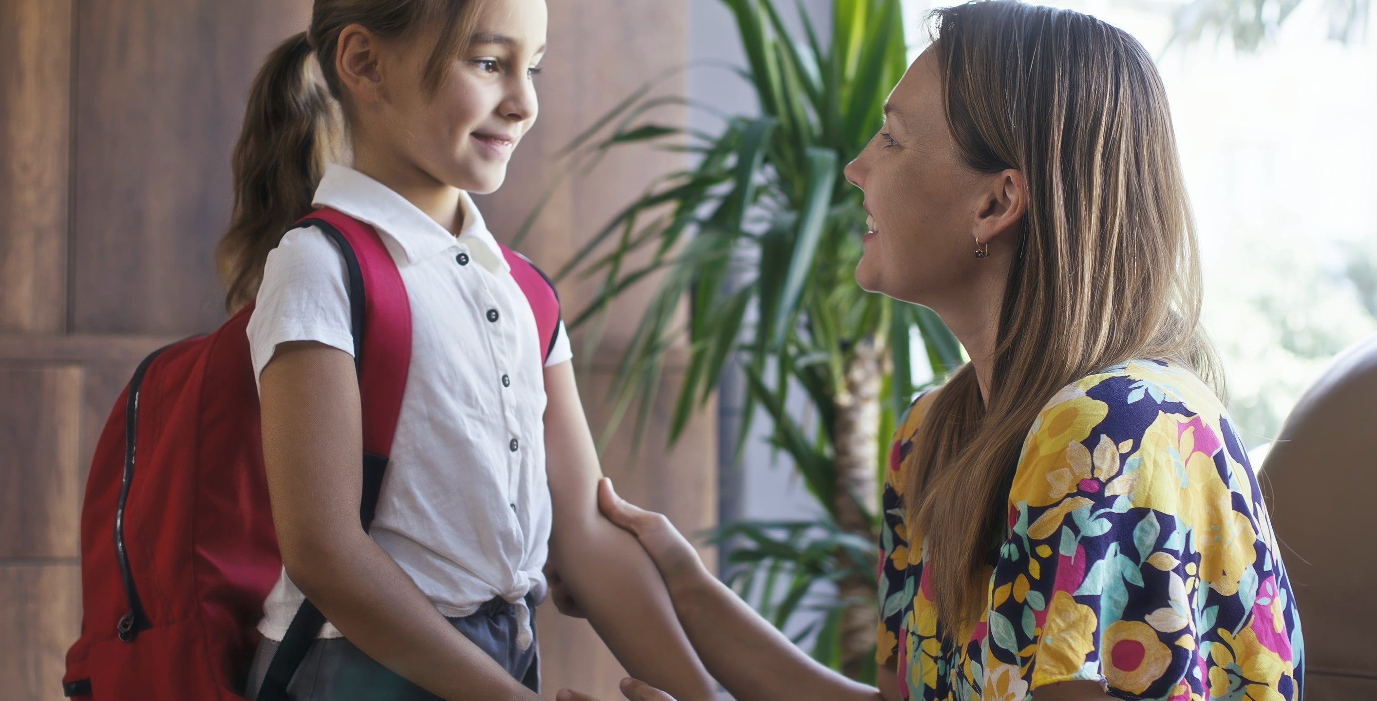 A young girl with a red backpack smiles at an adult woman in floral clothing who is holding her hands. They appear to be in a warm home setting