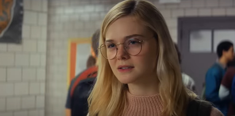 Elle Fanning wears round glasses and a casual outfit while standing in a hallway with other people in the background