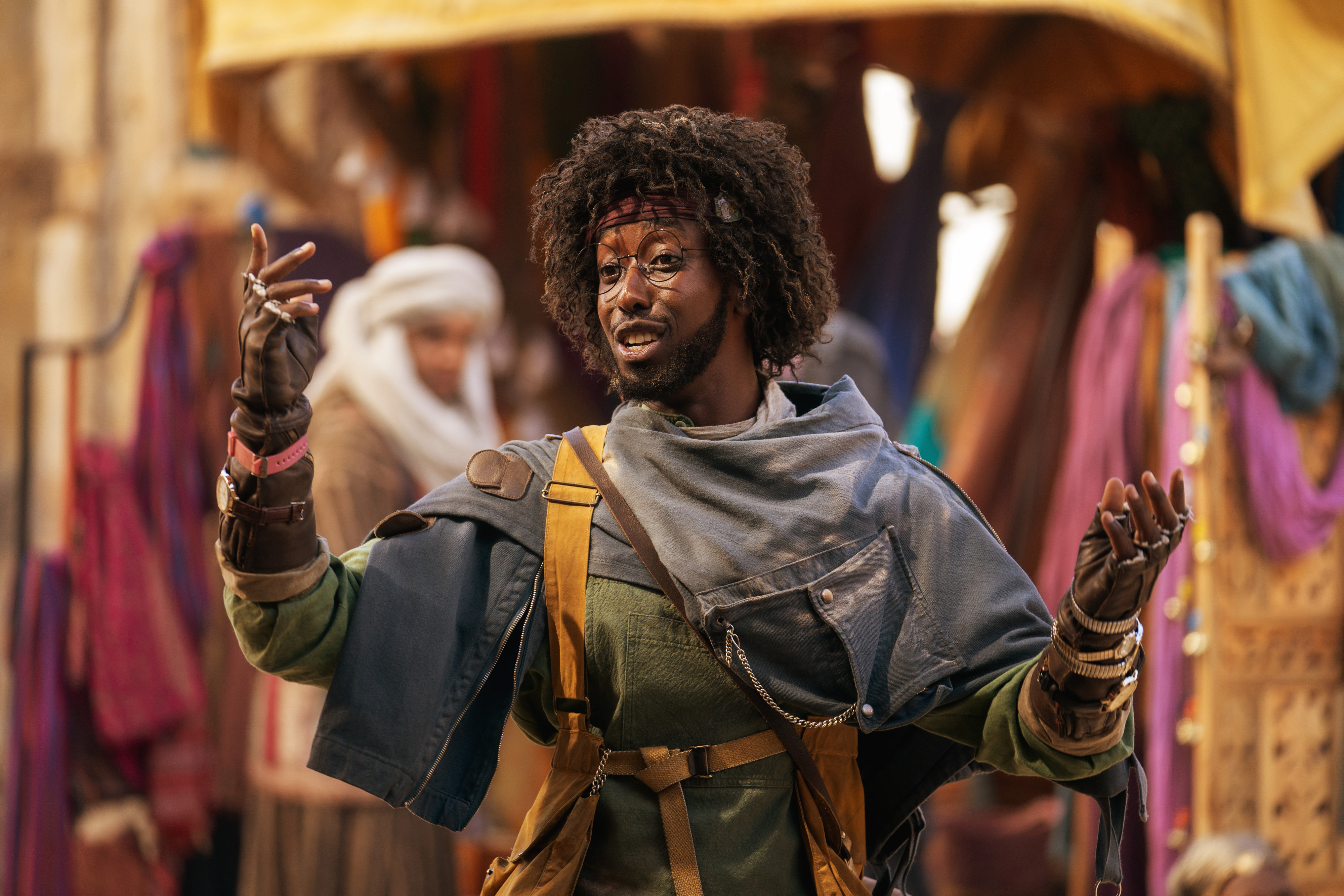 A person dressed in layered, adventure-style clothing, including a jacket and gloves, stands gesturing in a lively market scene