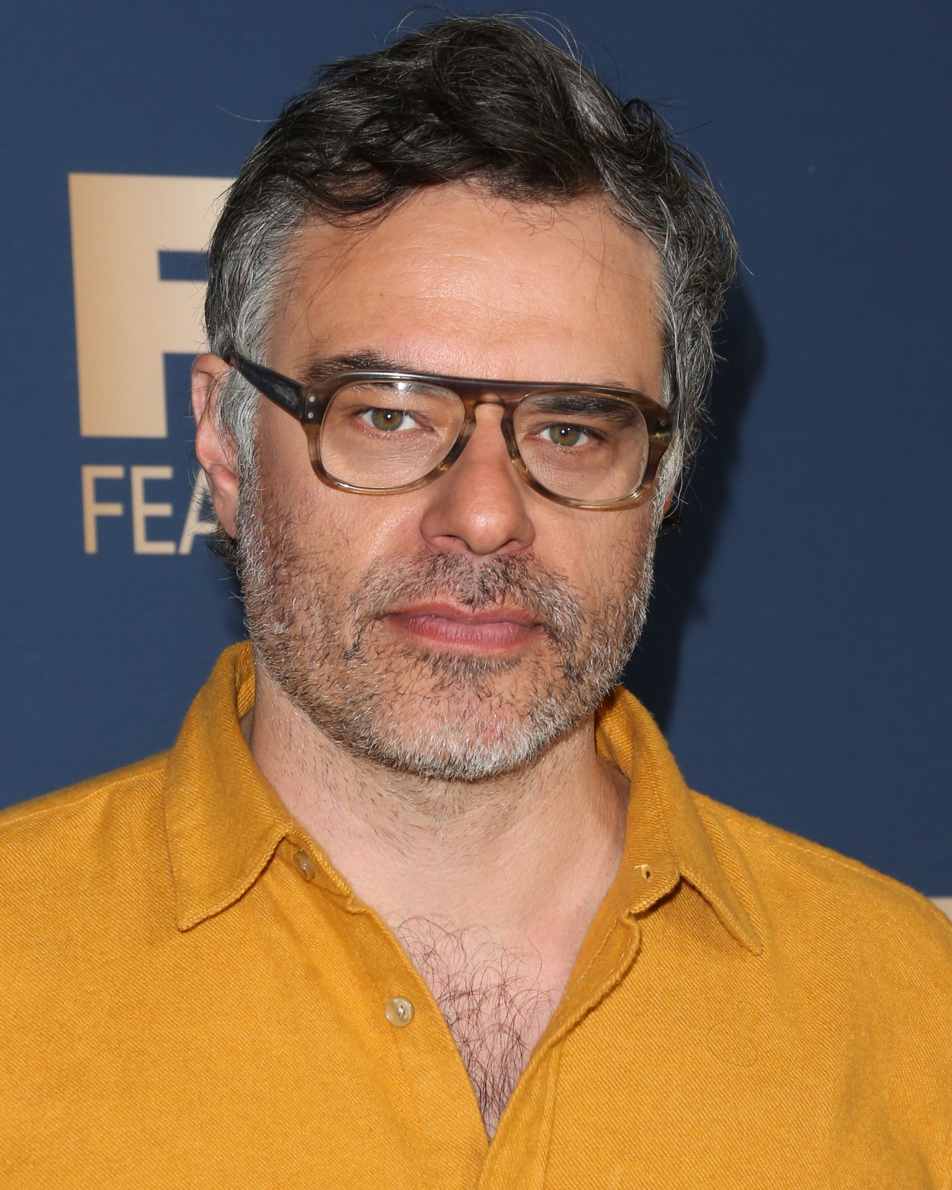 Jemaine Clement stands against a background, wearing a casual button-up shirt and glasses