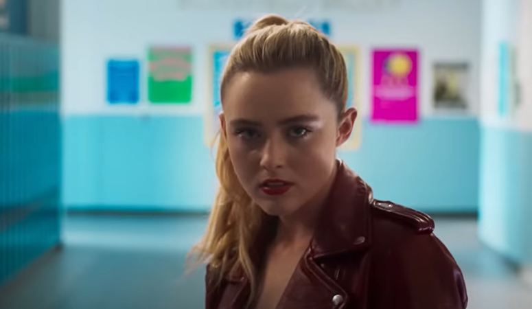 Kathryn Newton wearing a leather jacket, looking intently at the camera, with posters blurred in the background