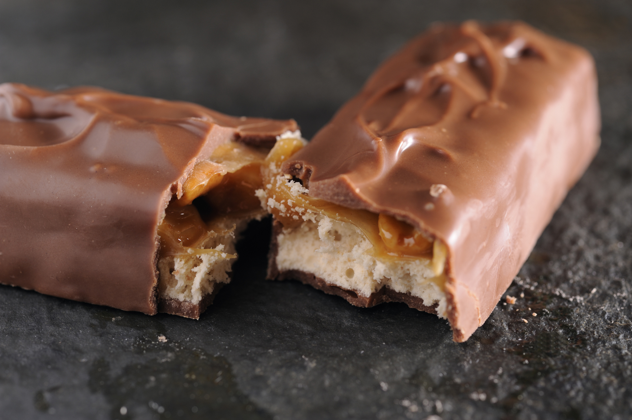 A broken chocolate-coated caramel and nougat candy bar sits on a black surface, showing its caramel and nougat insides