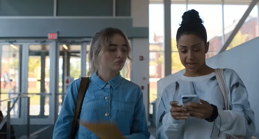 Sabrina Carpenter and Liza Koshy walk through a building. Sabrina holds a folder while Liza checks her phone and talks