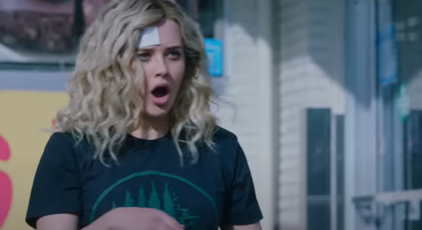 Katherine Langford with curly hair, a plaster on her forehead, and wearing a black T-shirt with a green tree print stands in front of a storefront, shocked.