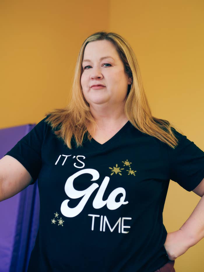 Woman wearing a black t-shirt that says "It's Glo Time." She has shoulder-length hair and is standing in front of a yellow background
