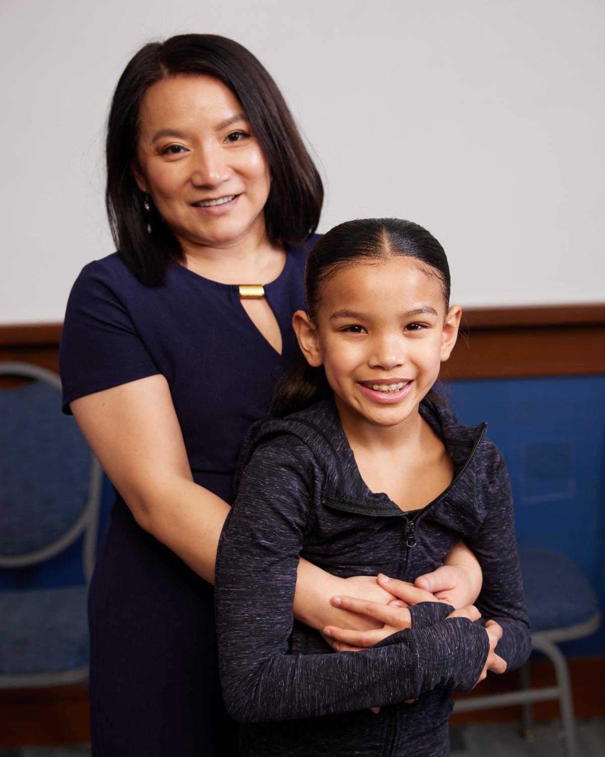 An adult and a young girl smile at the camera. The adult wears a short-sleeved dress, and the girl wears a zip-up jacket