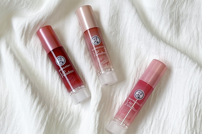 ベーシックコスメの新しいリップグロス3本が布の上に置かれています。シンプルでシックなデザインの広告用画像です。