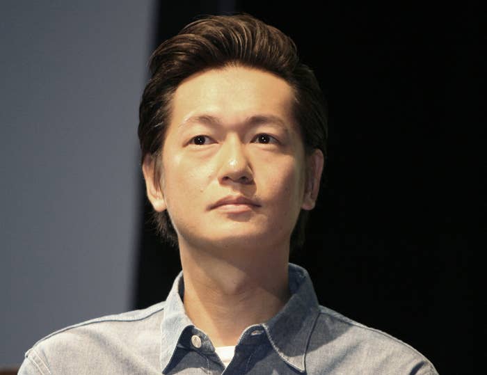 画像には男性が写っており、彼の名前は分かりません。デニムのシャツを着ています。背景には飲み物の瓶があります。