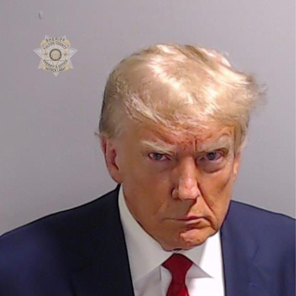 Donald Trump in a suit and tie, facing forward for a mugshot with a stern expression. Sheriff's star badge is in the background