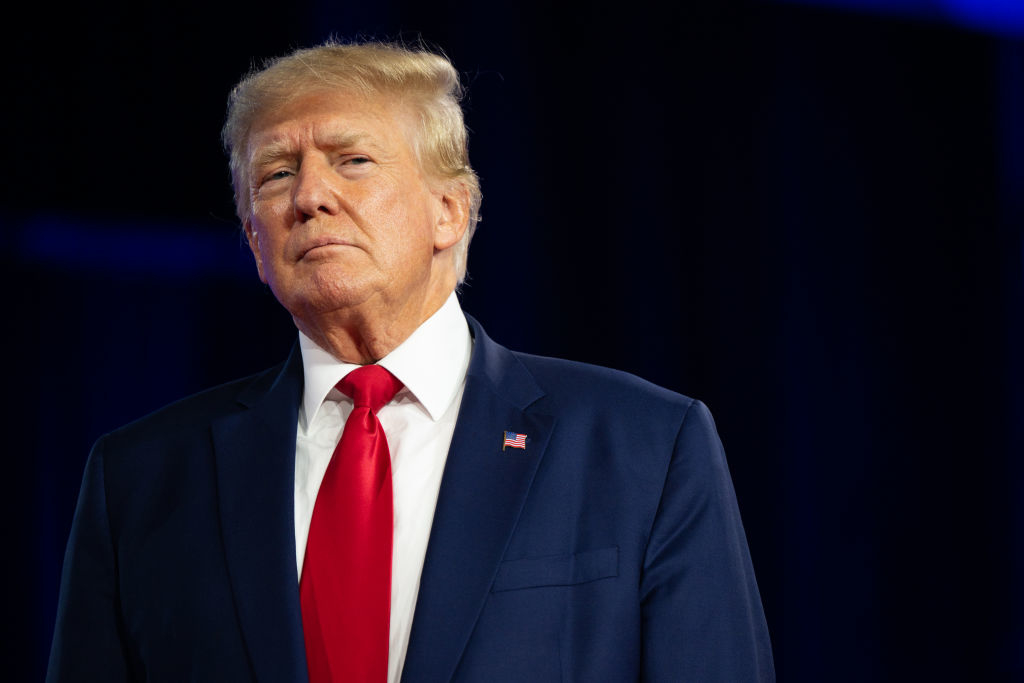 Donald Trump wearing a suit with a red tie, standing against a dark background