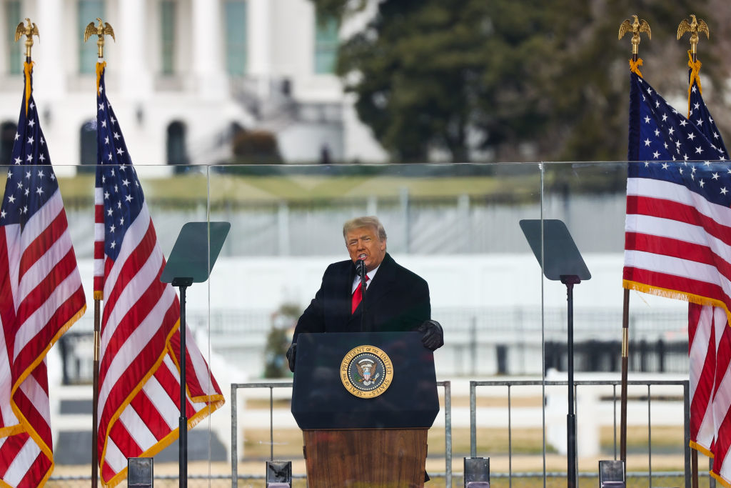 Donald Trump speaking at a podium with the Presidential Seal, outdoors, flanked by U.S. flags, with the White House in the background