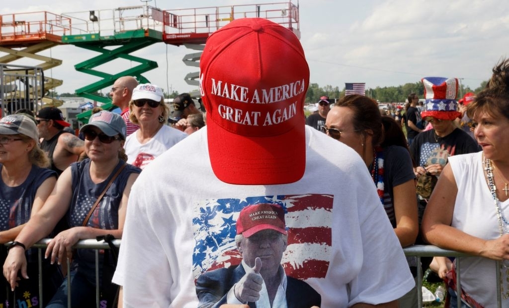A person wearing a red "Make America Great Again" hat and a T-shirt with an image of Donald Trump. They are standing in a crowd at what appears to be an outdoor event