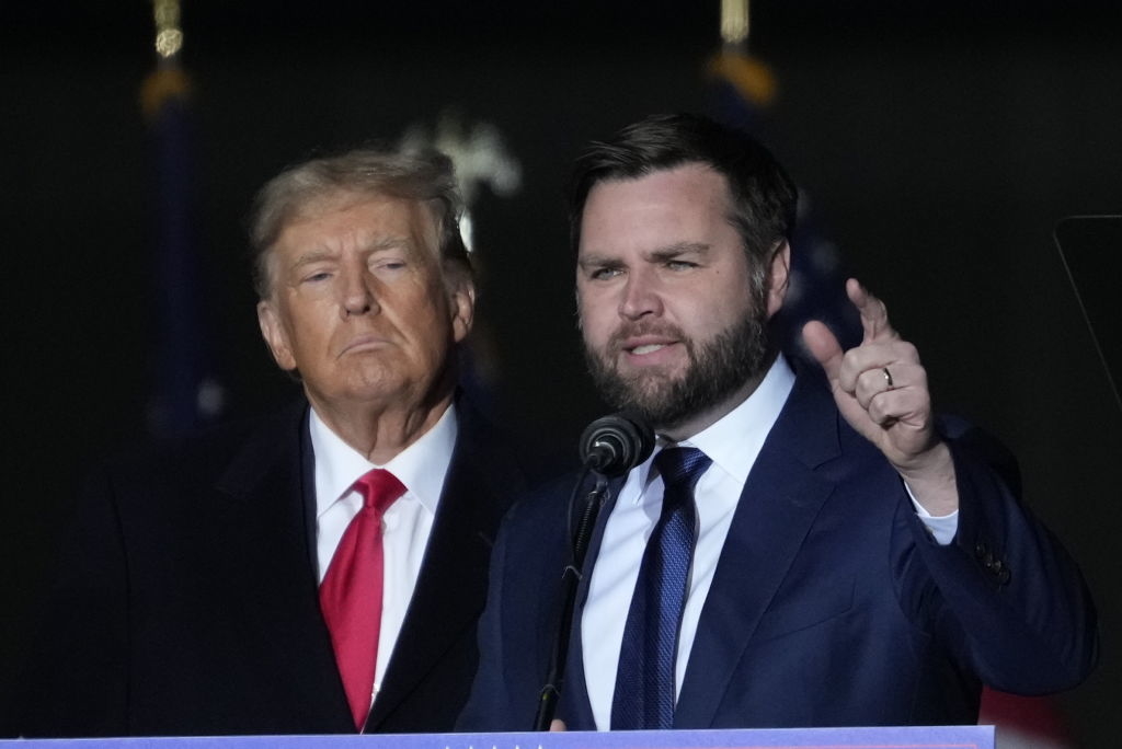 Donald Trump in a suit with a red tie stands next to J.D. Vance in a suit and blue tie, as Vance speaks on stage at an event
