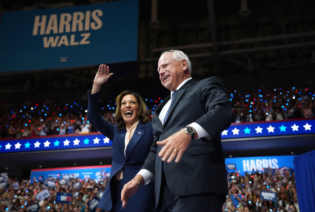 Kamala Harris and Tim Walz wave to a cheering crowd at a rally. A large sign behind them reads "Harris Walz."