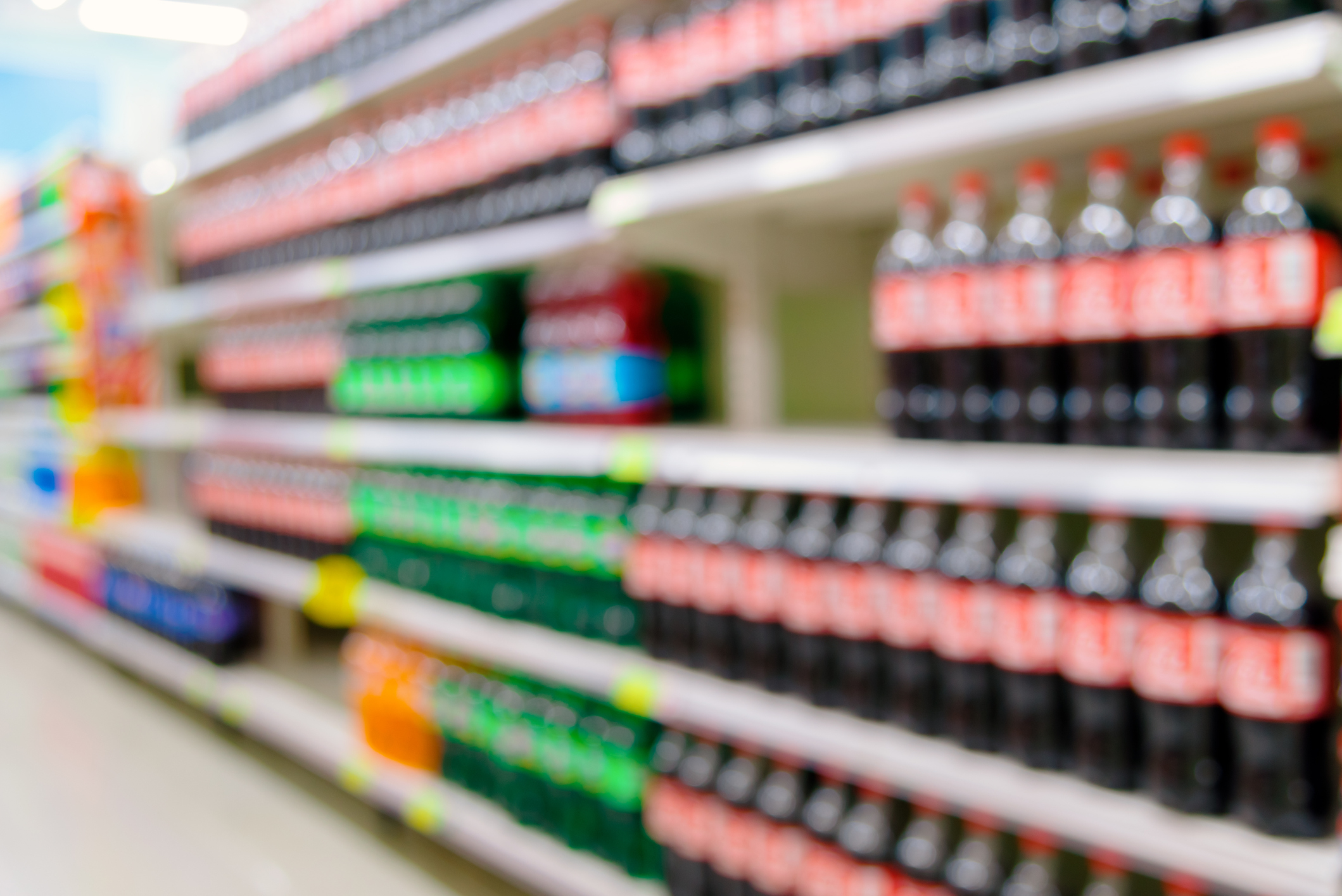 A blurry image of supermarket shelves stocked with various soda bottles, with Coca-Cola bottles prominently visible
